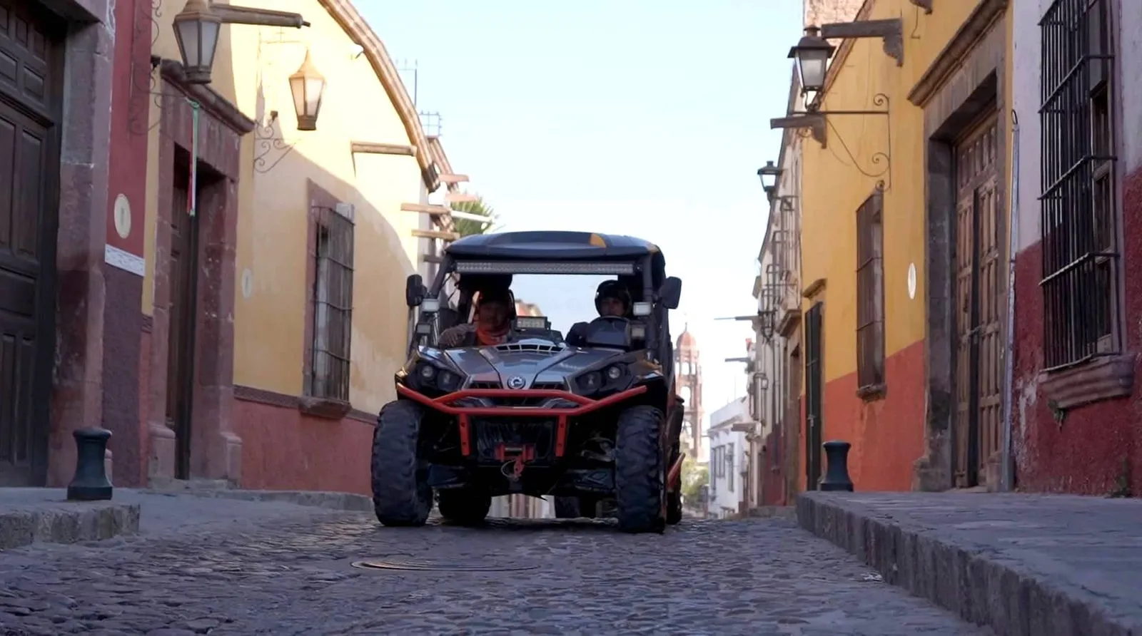 Touring the historic cobblestone streets of Mexico with Rzr rentals. Front view of a gray UTV Rzr rentals vehicle driving up a narrow cobblestone street flanked by colorful pastel colonial buildings in a historic Mexican town.
