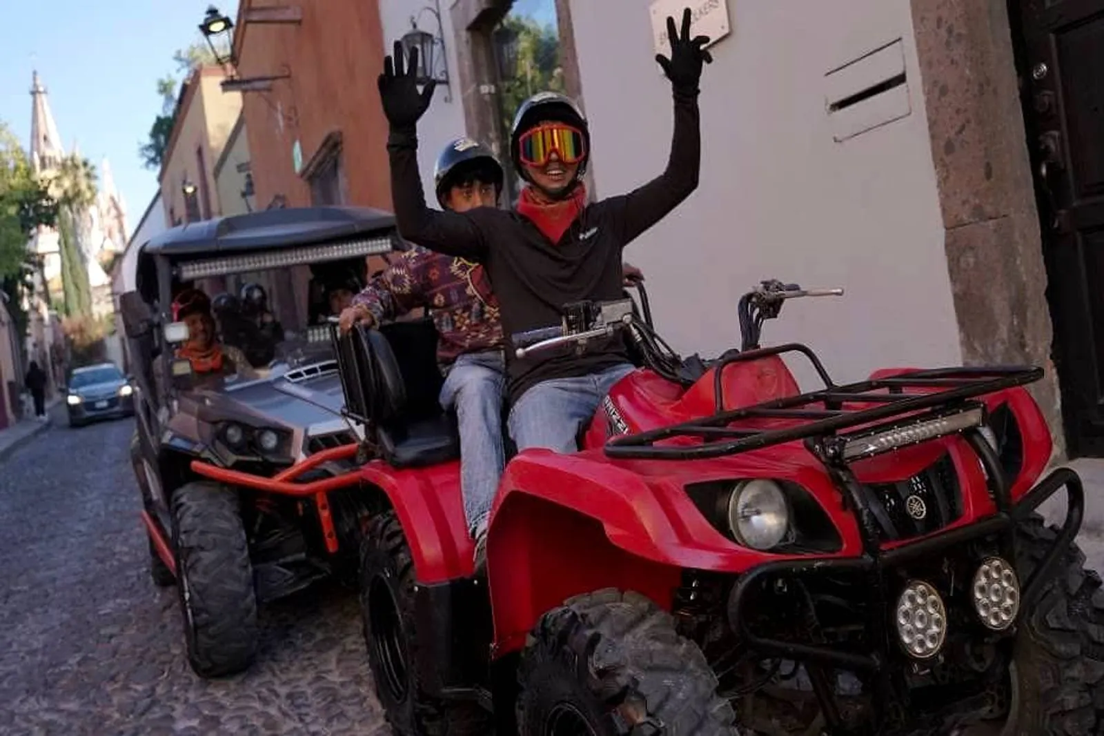 Family adventure experience with Rzr rentals and ATV riding. A happy child wearing a helmet and colorful goggles on a red ATV, smiling and waving both hands at the camera while riding behind a gray Rzr rentals vehicle on a colonial street.